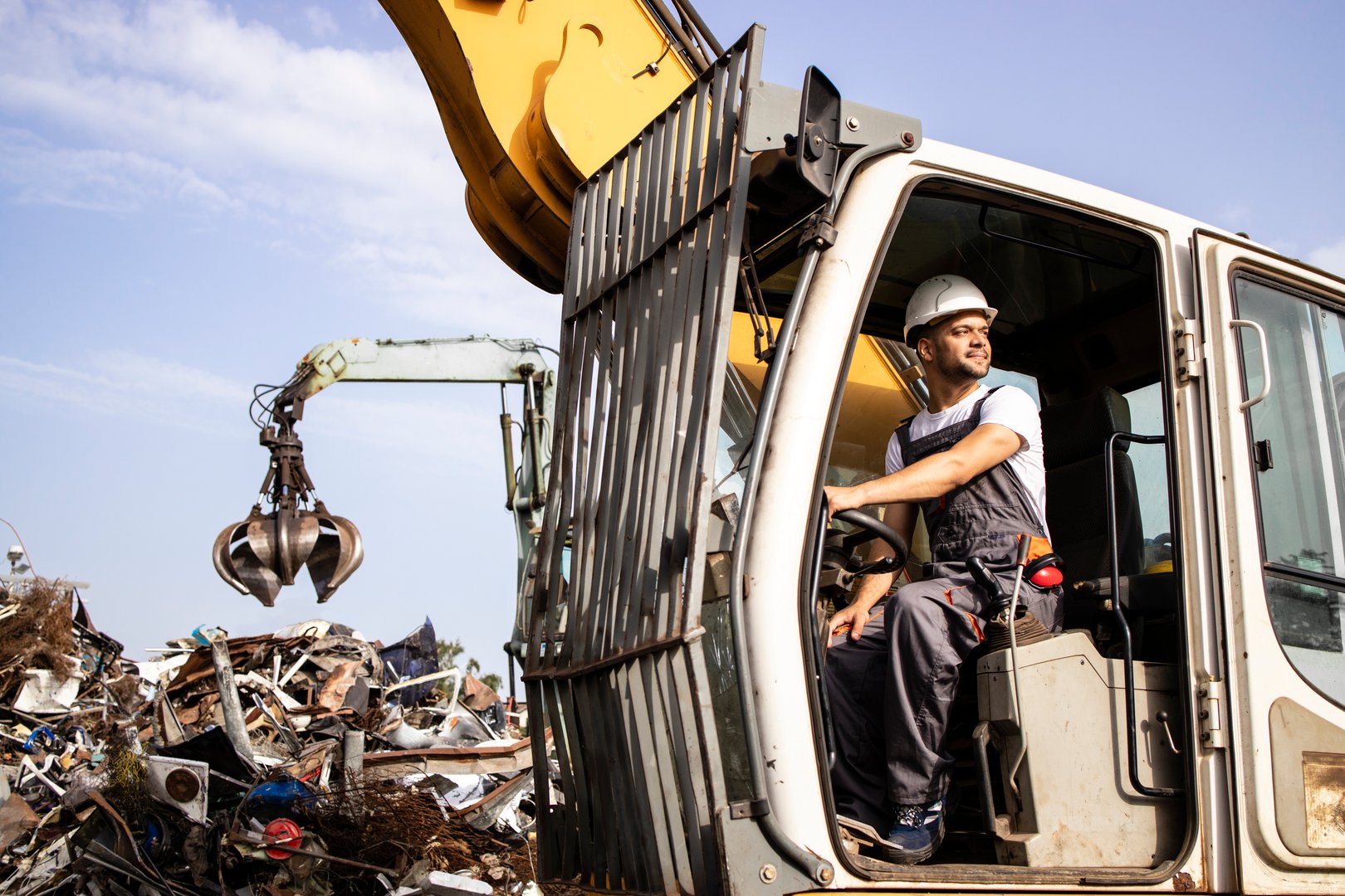 Man operating excavator industrial machine with claw attachment used for lifting scrap metal in junk yard.