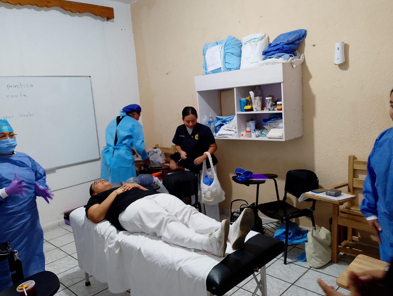 Medical training session with a patient on a table, surrounded by healthcare workers in scrubs and protective gear.