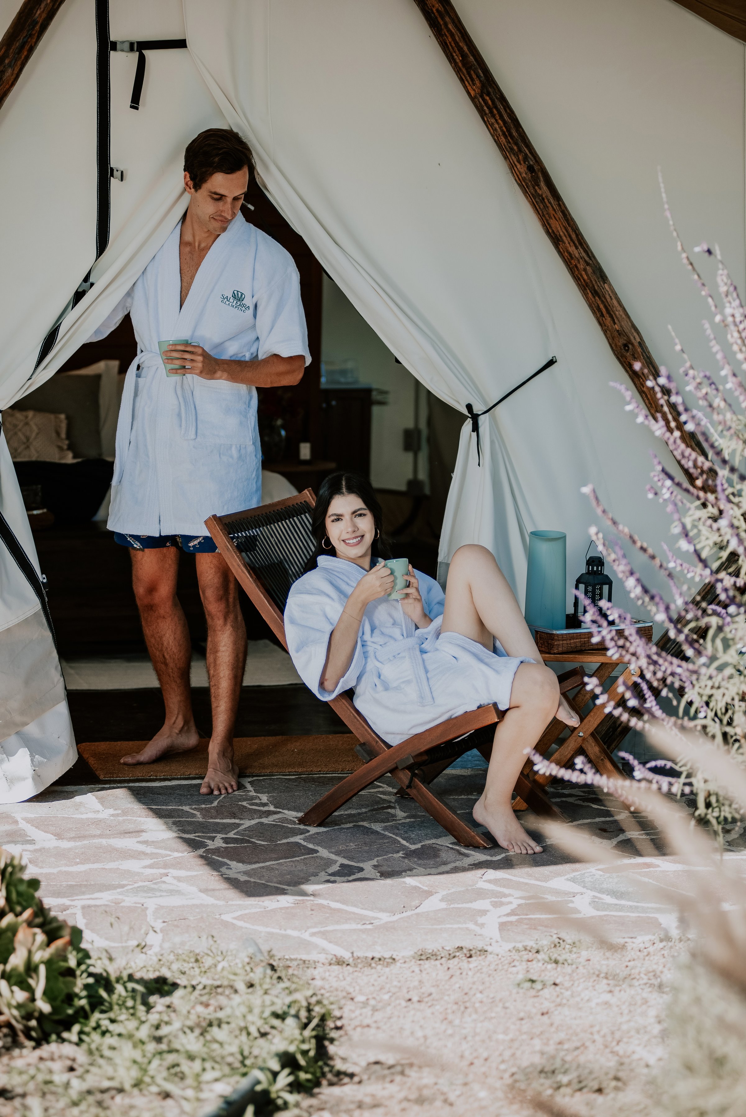 Couple in robes relaxing outside a glamping tent, holding drinks, with lavender plants nearby, on a sunny day.