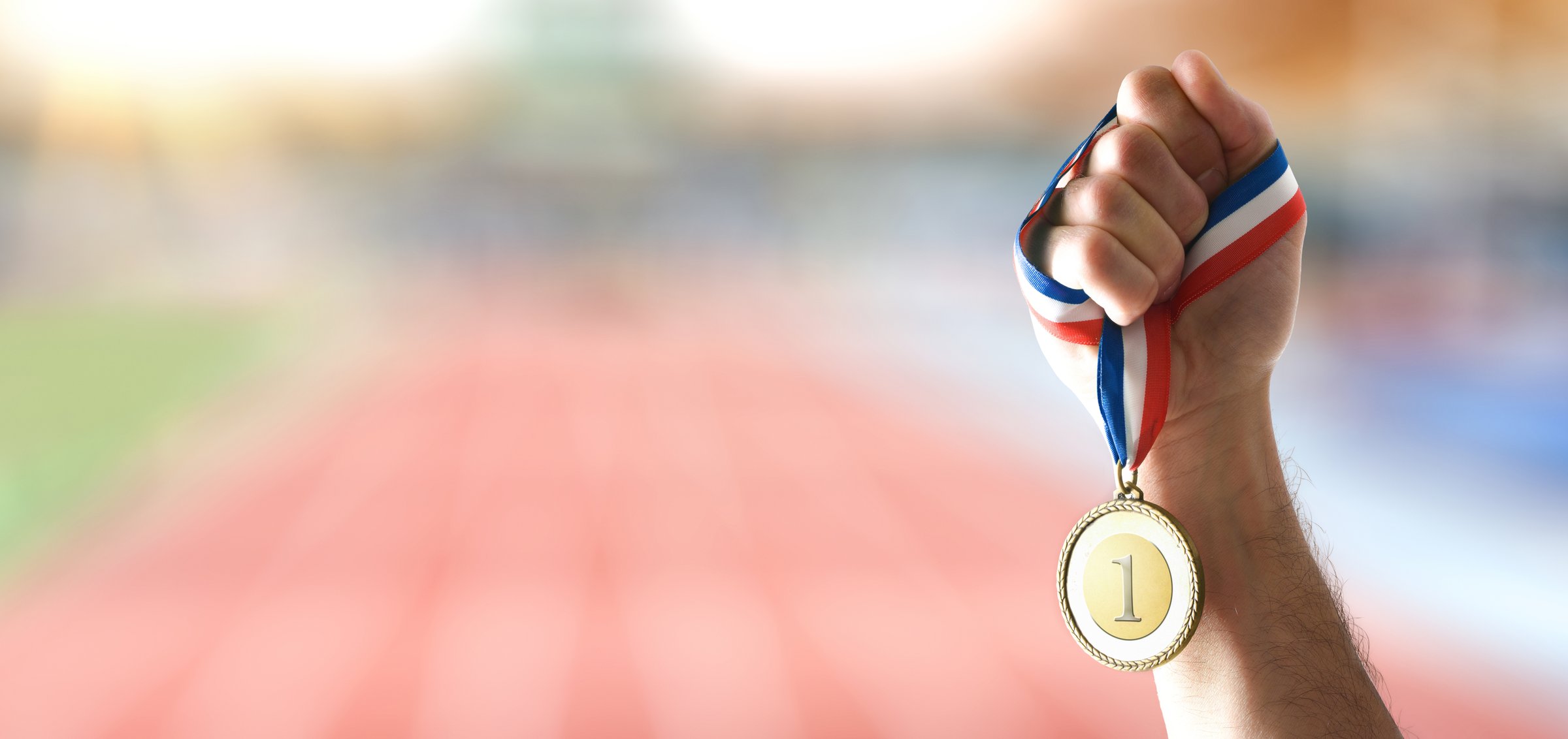 Hand showing gold medal with engraved number 1 and athletics stadium behind a sunny day.