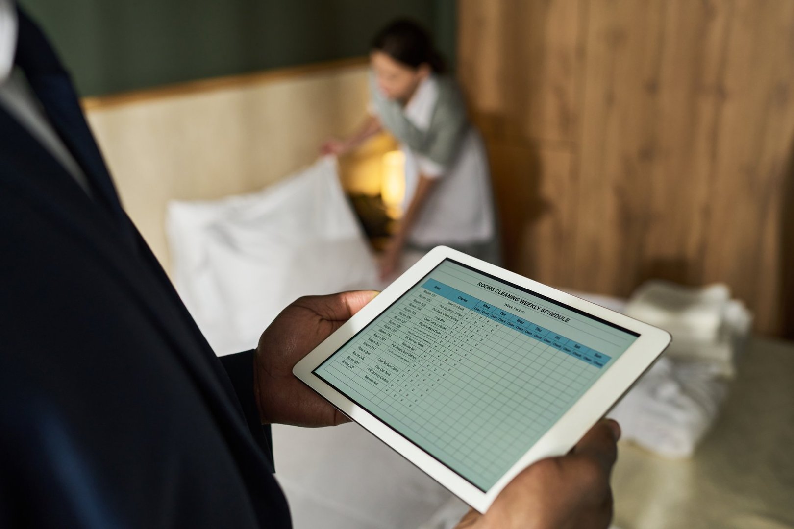 Business person managing hotel staff using tablet technology for housekeeping operations in hotel room setting while focusing on digital data and lists of duties