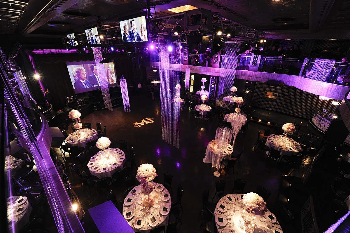 Elegant event venue with round tables, floral centerpieces, chandeliers, and video screens showing people. Ambient purple lighting.