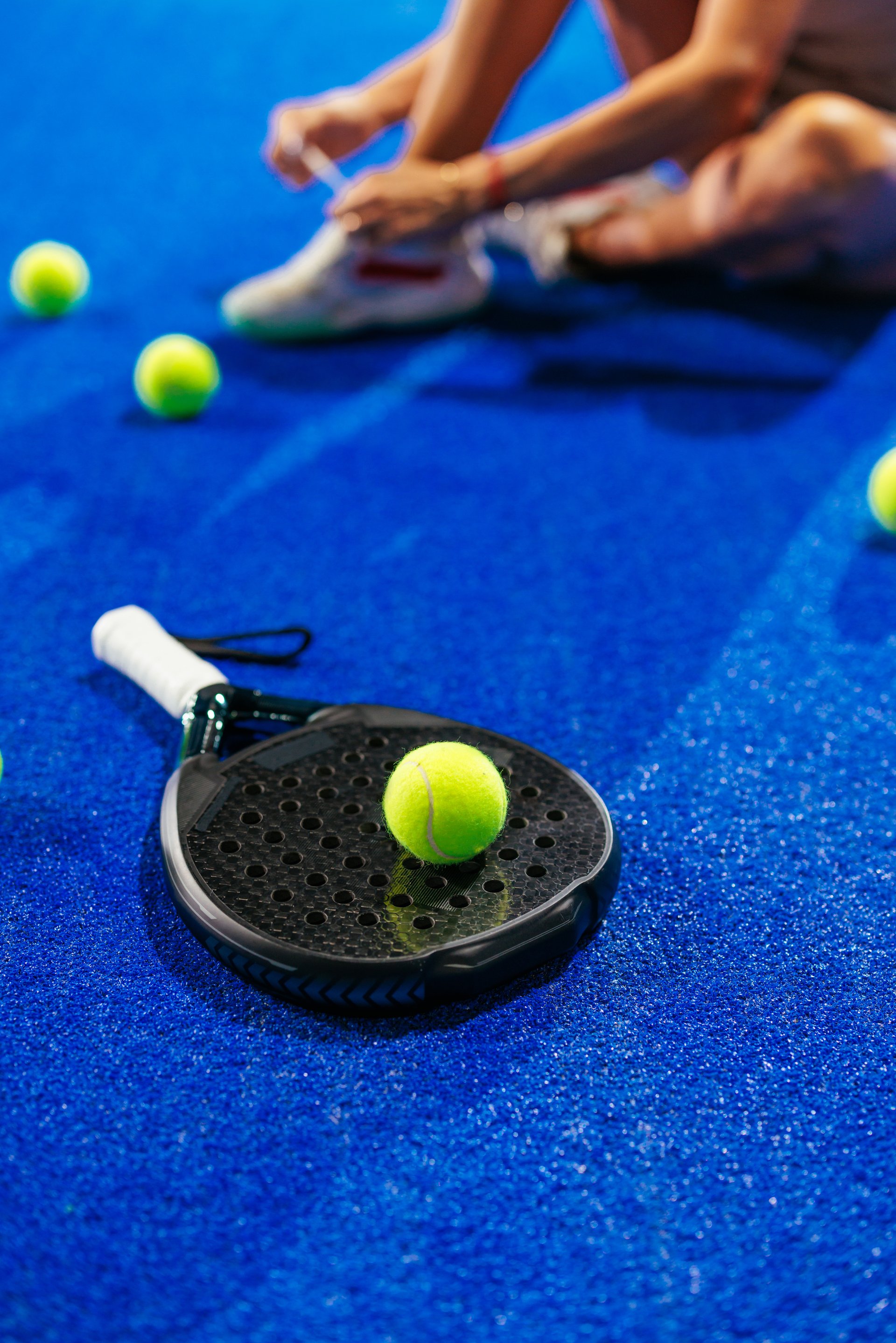 Close up of a padel racket and padel ball on top of it on a blue surface of a padel court with a person sitting and tying shoelaces blurred in the background.