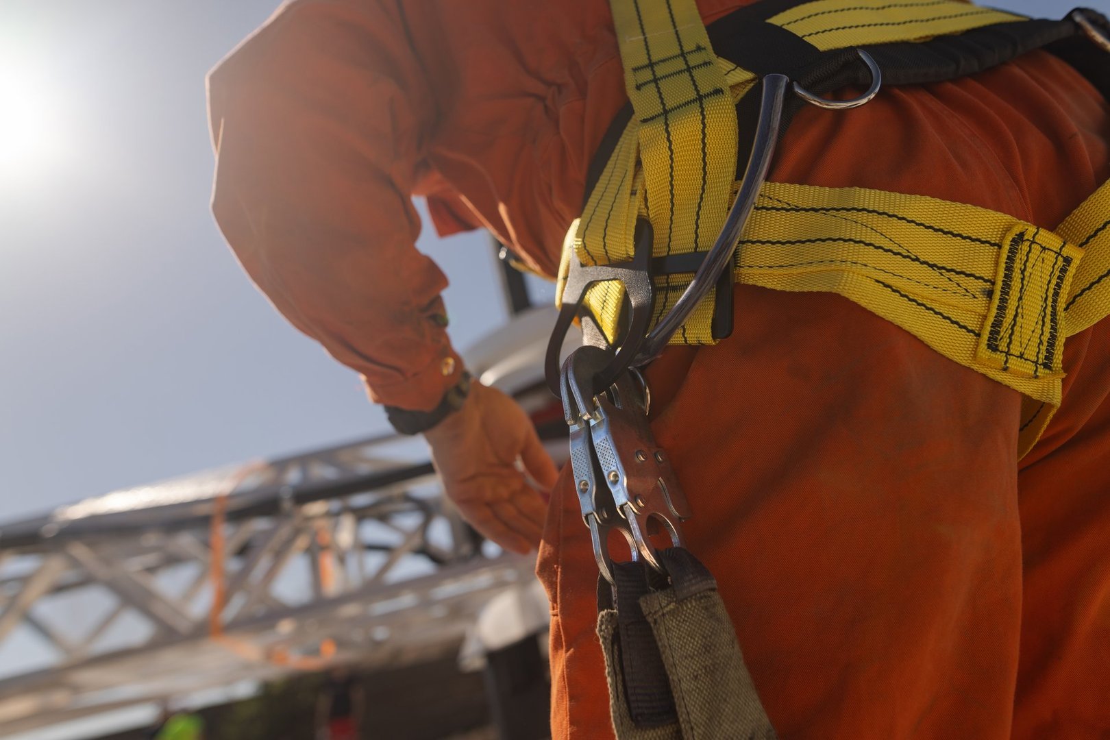 A close-up of a worker wearing a safety harness with a yellow strap, preparing to climb or work at height. The background shows a construction site with bright sunlight.