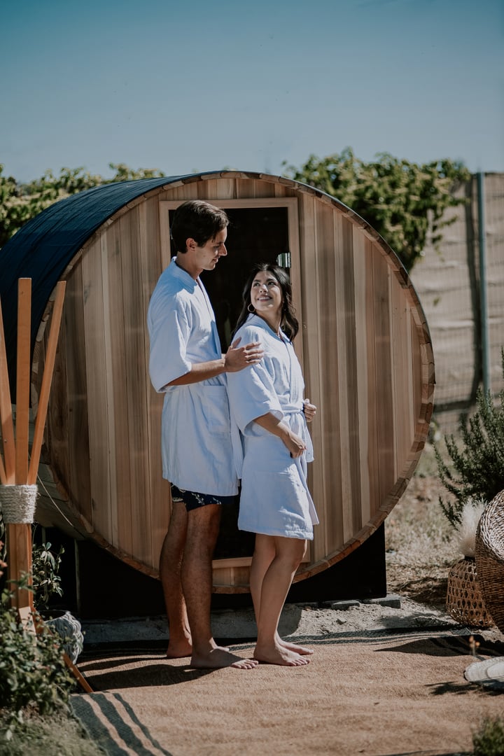 A couple in matching robes stands smiling outside a wooden sauna barrel on a sunny day.