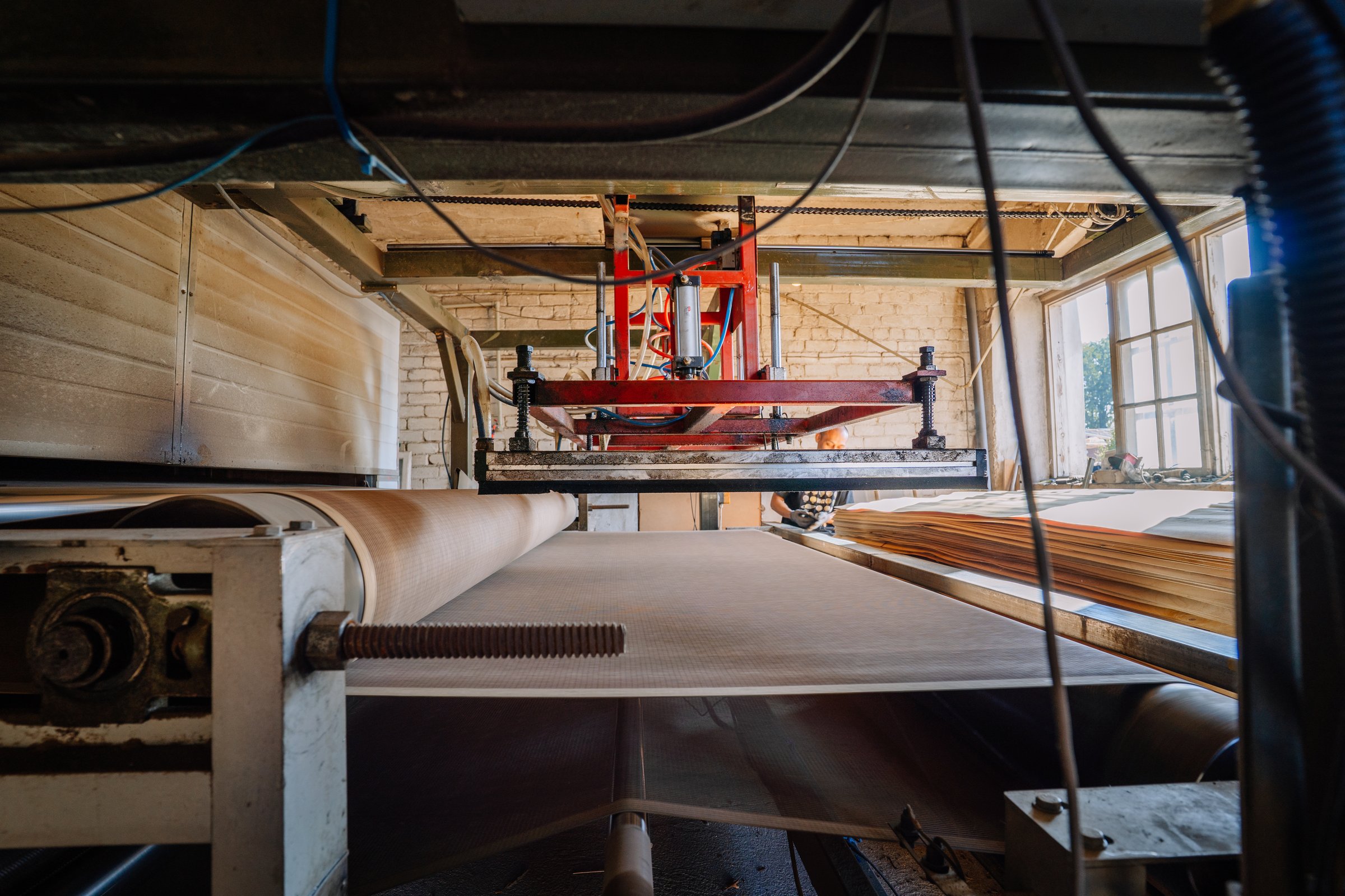 Valmiera, Latvia - September 10, 2025 - A plywood factory machine processes veneer sheets on a conveyor belt as a worker in the background oversees the production under natural light.