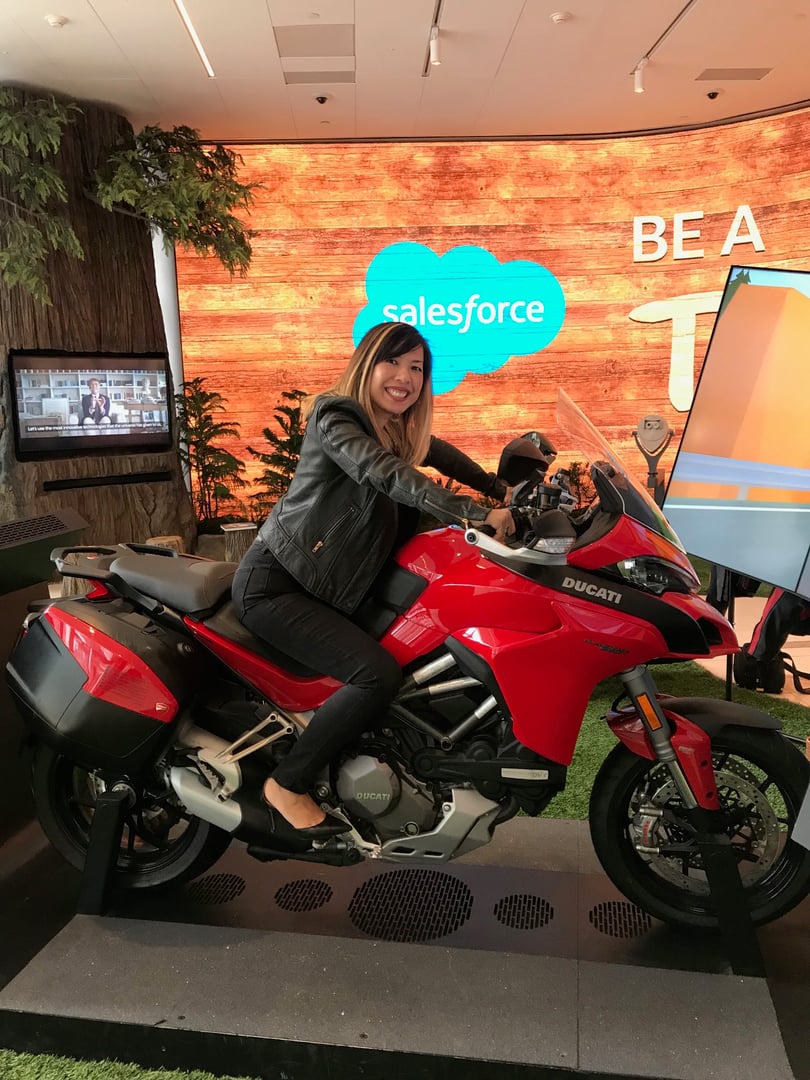 Smiling person sitting on a red Ducati motorcycle in a showroom with a Salesforce sign in the background.