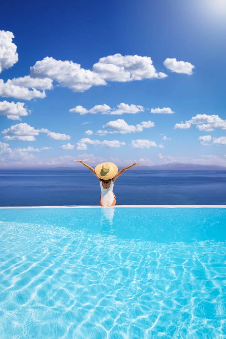 Summer holiday concept with a happy woman with sunhat sitting at the edge of a big infinity pool with view over the ocean