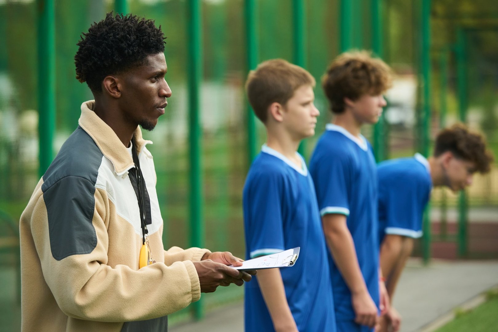 Black man holding clipboard coaching group of teenage boys standing in line on sports field during outdoor team practice session