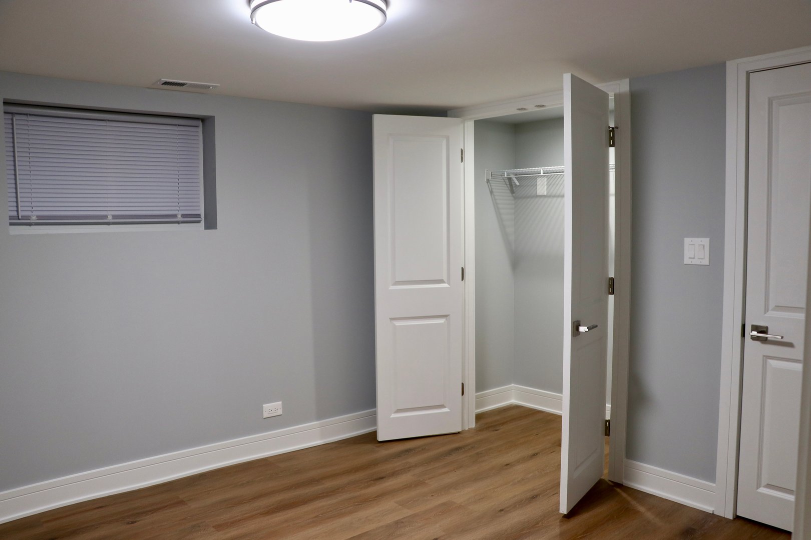 Empty room with a wooden floor, light gray walls, a small window, and an open closet with white sliding doors.