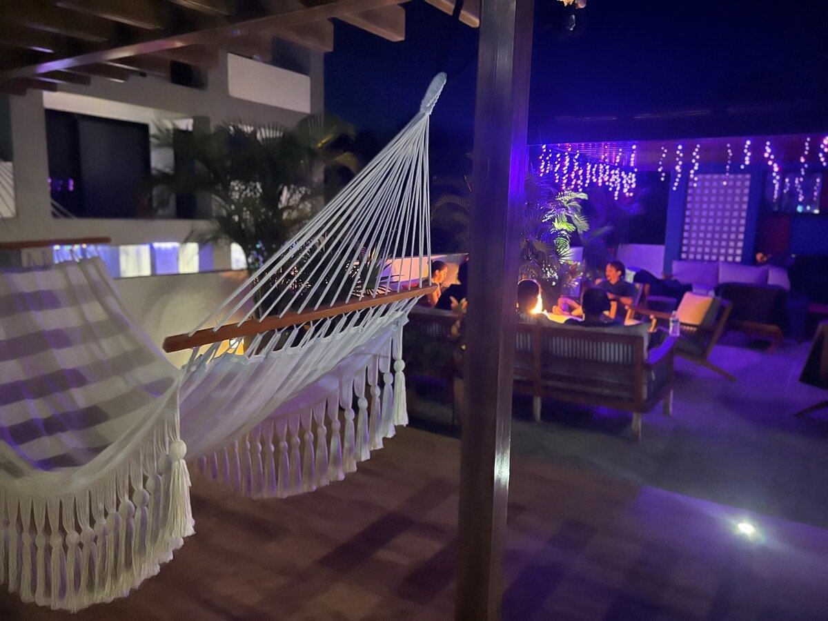 Outdoor lounge area at night with a white hammock and people sitting on sofas under purple lights.