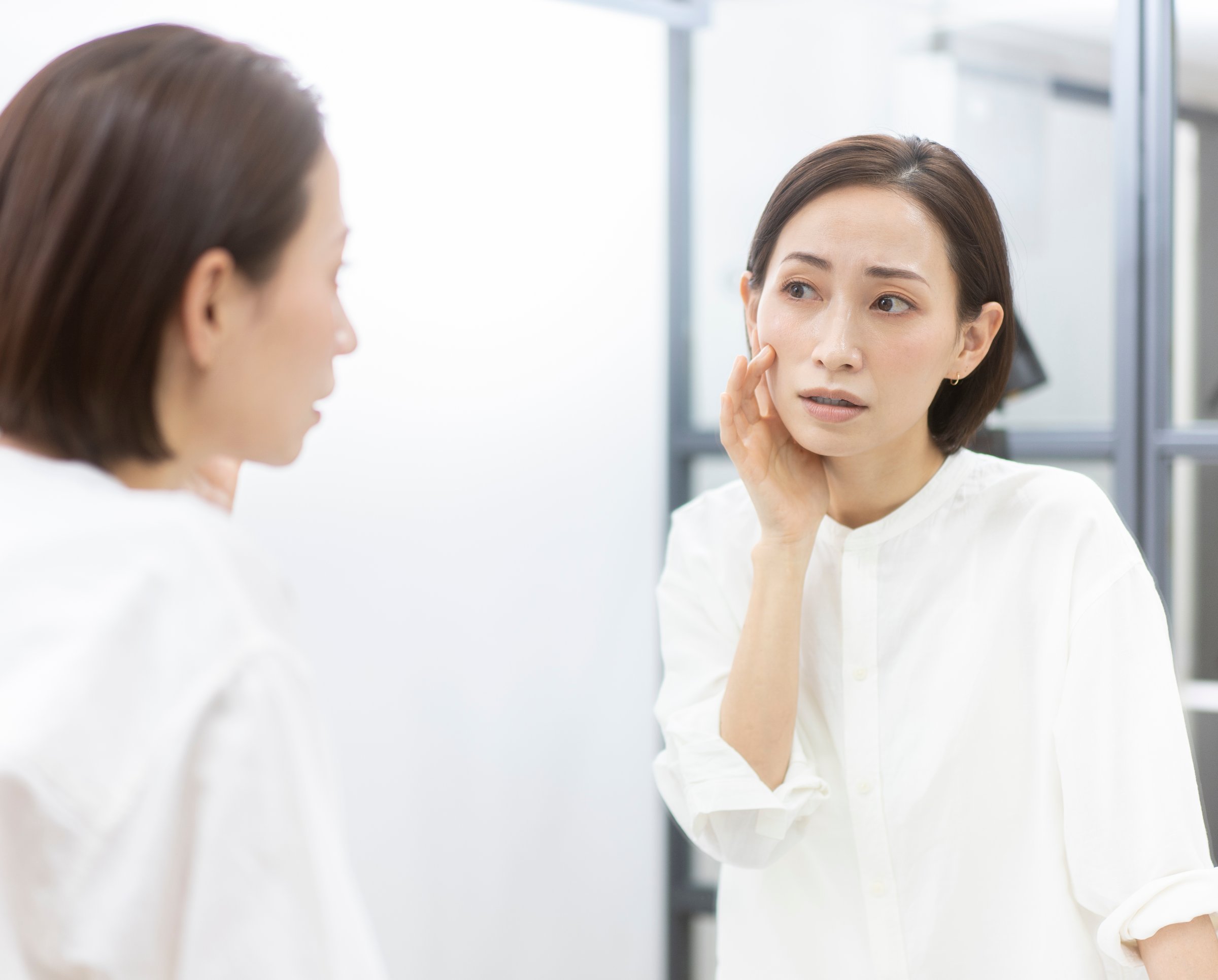 A middle-aged Japanese woman is touching her skin while looking in the mirror.