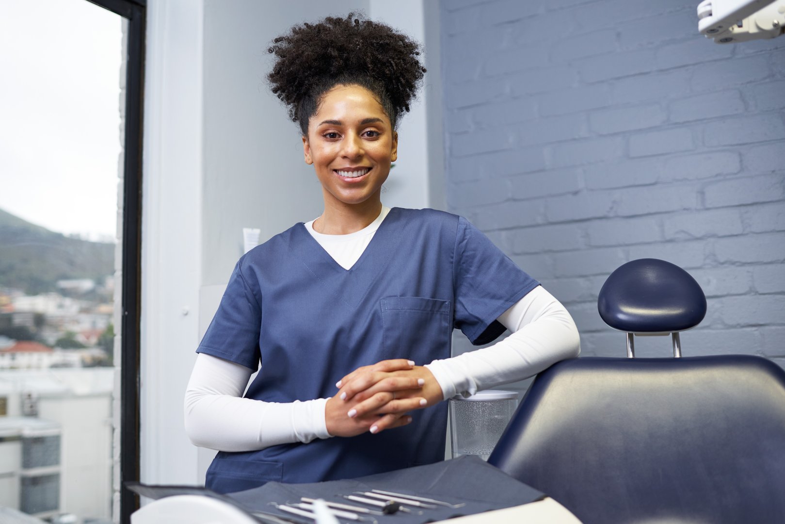 Confident dentist posing in modern clinic, wearing navy blue scrubs. Professional healthcare setting with dental chair, tools, and bright interior, reflecting expertise, patient care, and dedication in dentistry.