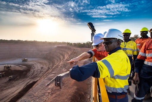 Mining engineers in safety gear examining an open-pit mine at sunrise, discussing operations, with machinery in the background.