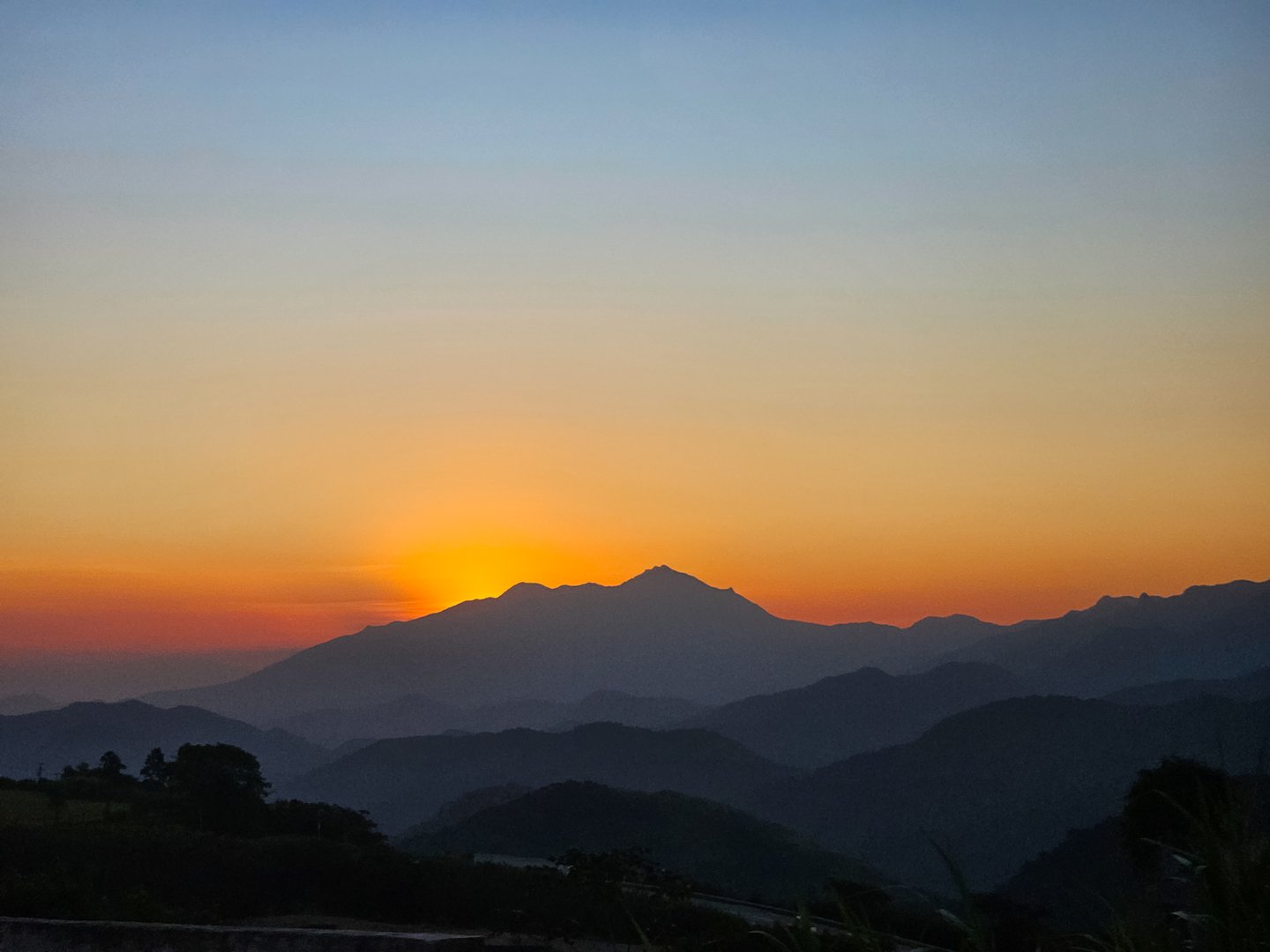 Sunrise over the mountainous region of Petrópolis (Brazil), with the silhouette of mountains in the background. The sky transitions from deep blue at the top to a warm orange glow near the horizon, casting light over the layers of overlapping hills and valleys.