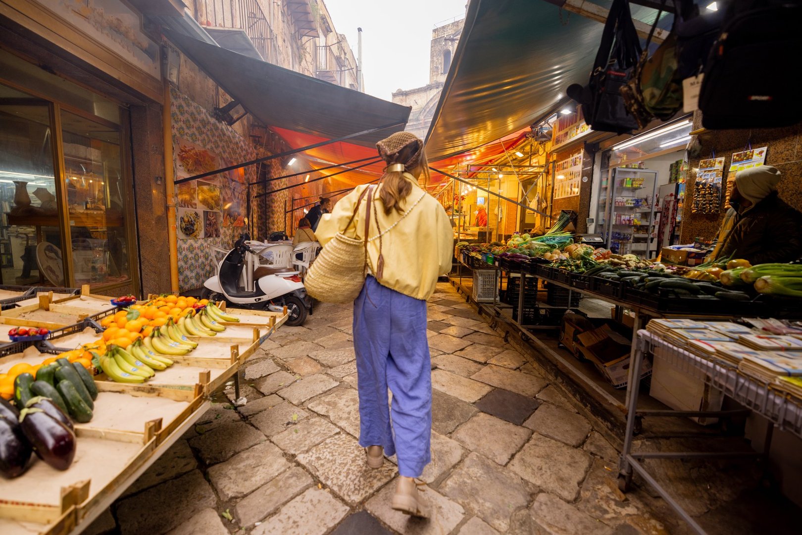 A woman in a yellow jacket browses fresh lettuce at an open-air vegetable market in Sicily, holding her phone. This image represents sustainable living, mindful food choices, and local tourism appeal