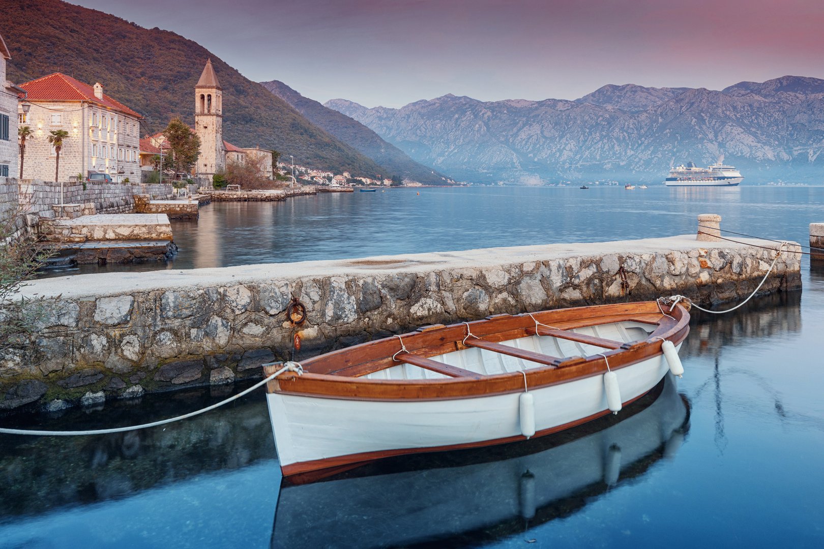 Wooden boat moored at the dock in coastal village Donji Stoliv, Montenegro, with a cruise ship in the background during sunrise