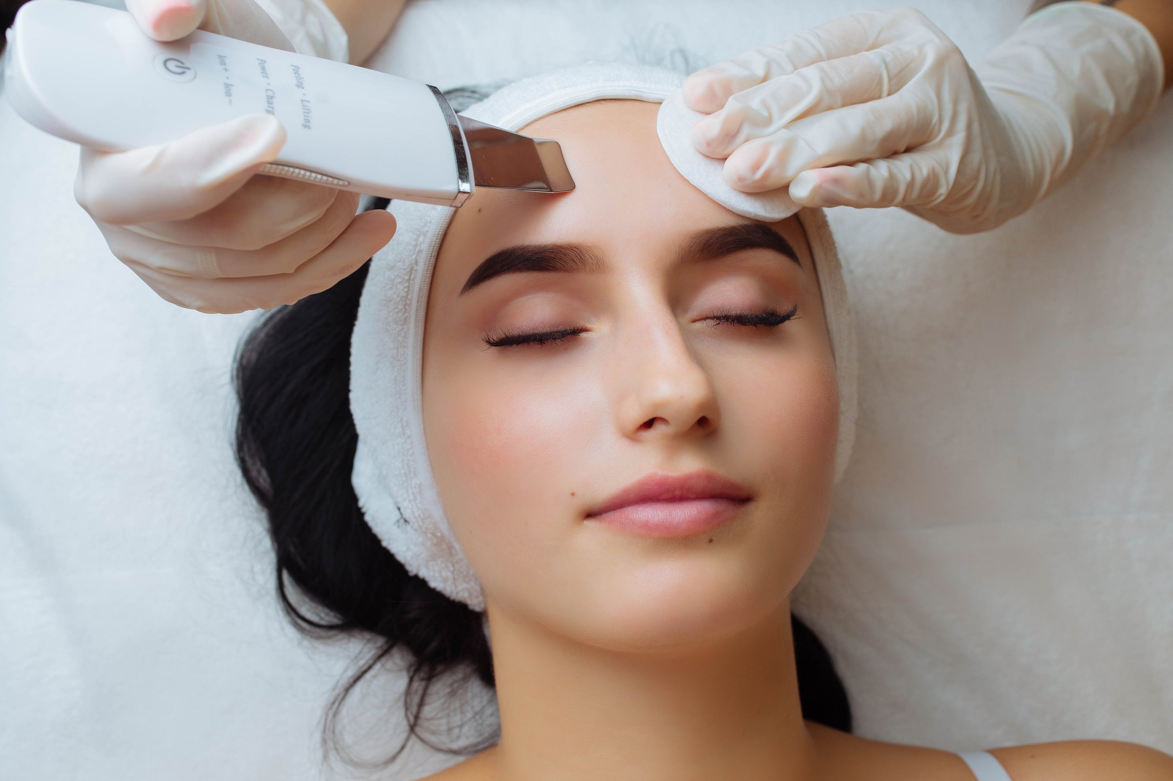 Close up top view of pretty young Caucasian lady during the procedure of cleansing the skin with ultrasound scrubber. Ultrasonic device in the hands of professional female afro beautician