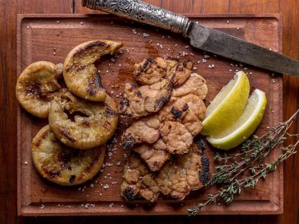 Grilled mushrooms and sausages on a wooden board with lemon wedges, thyme sprigs, and a rustic knife.
