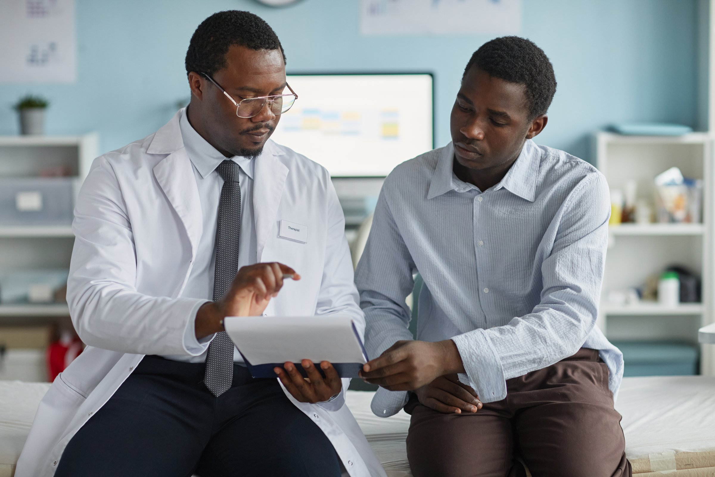 Black middle aged male doctor explaining medical information on digital tablet to Black young adult man patient during consultation in modern medical office