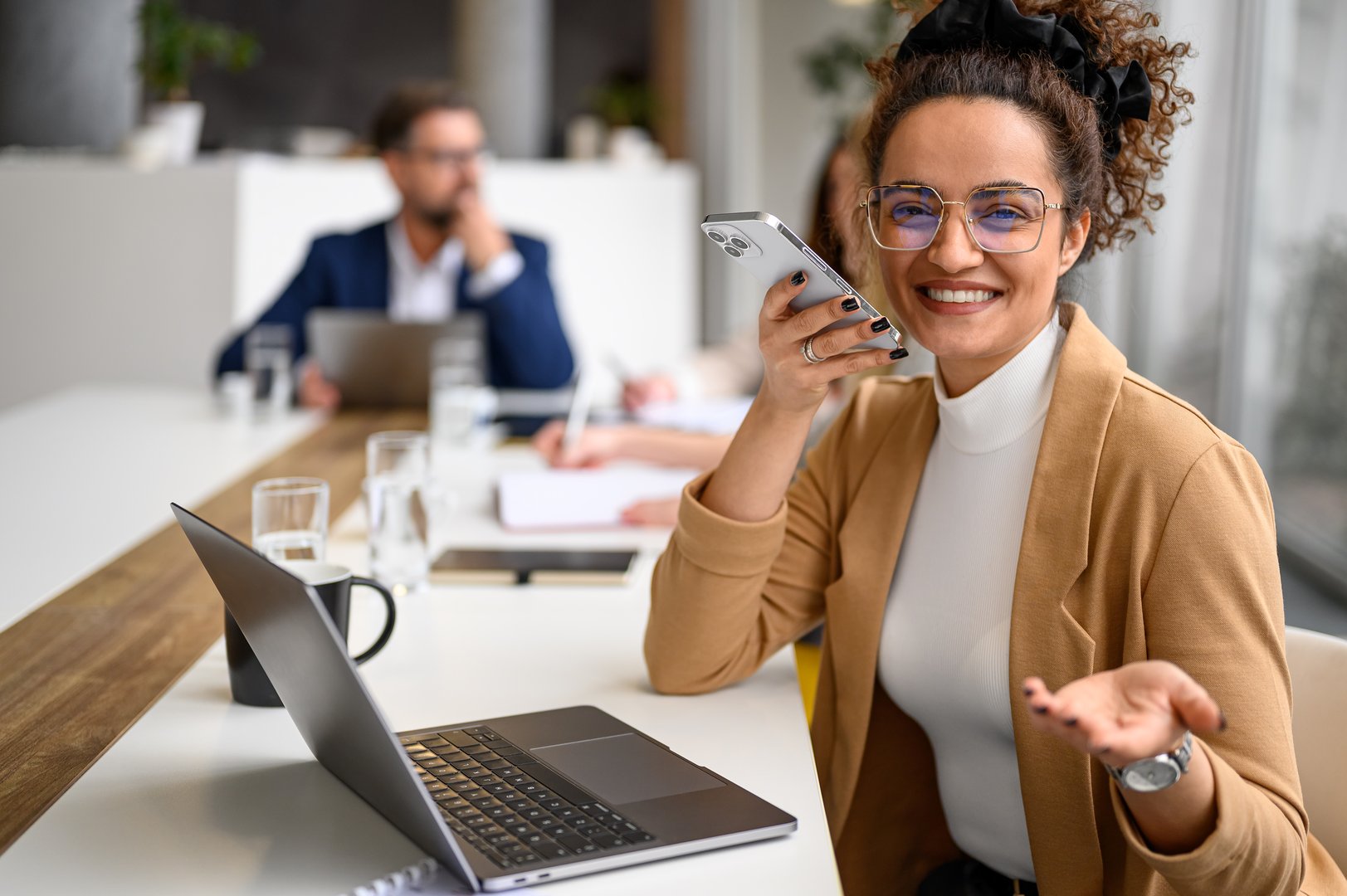 Happy manager in glasses speaking on her mobile phone's speaker while business coworkers collaborate in the background.