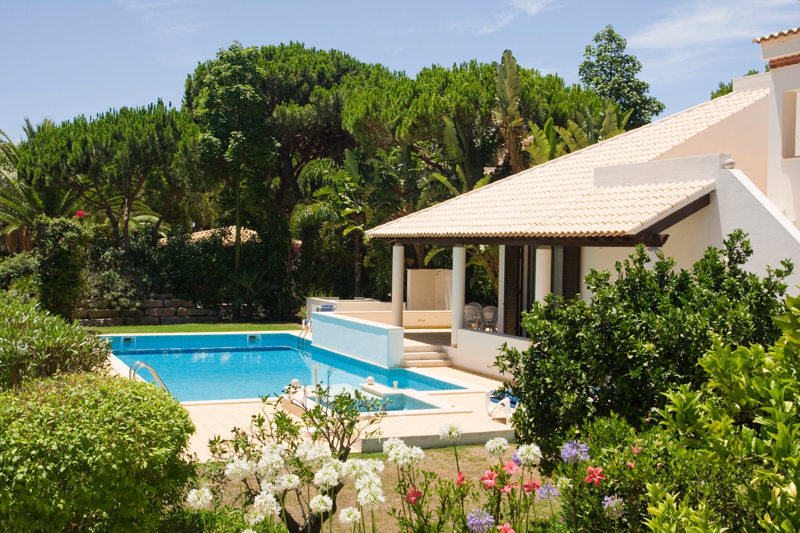 Luxury villa with a pool, surrounded by lush greenery and flowers, featuring a shaded patio area.
