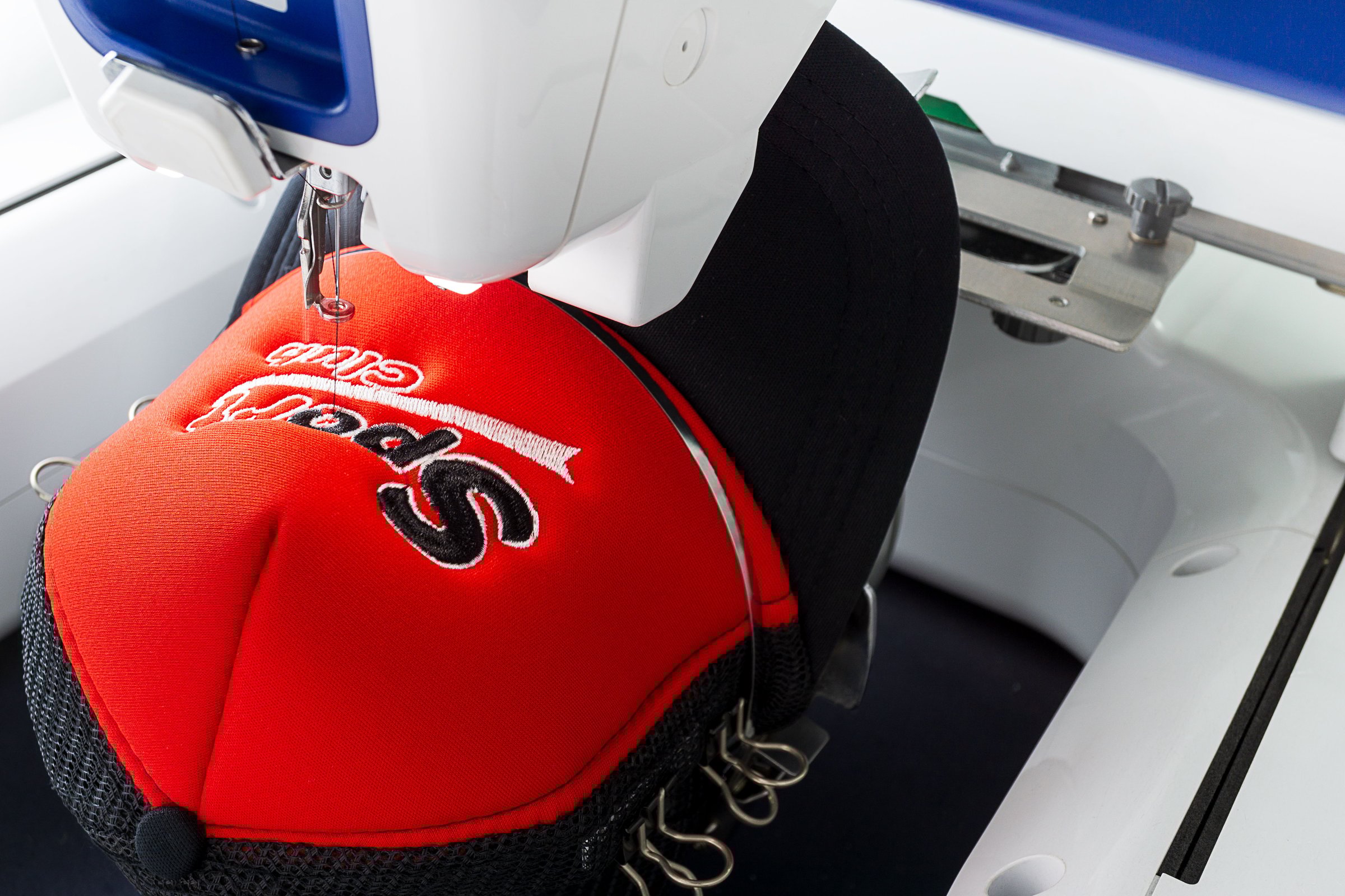 Close up image of white embroidery machine embroidering logo on red and black sport cap