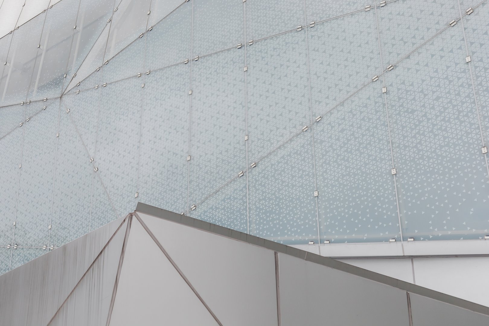 A modern building showcases a glass facade with a geometric pattern. The design includes metal angles and reflects light during the day. The scene emphasizes architecture and structure.