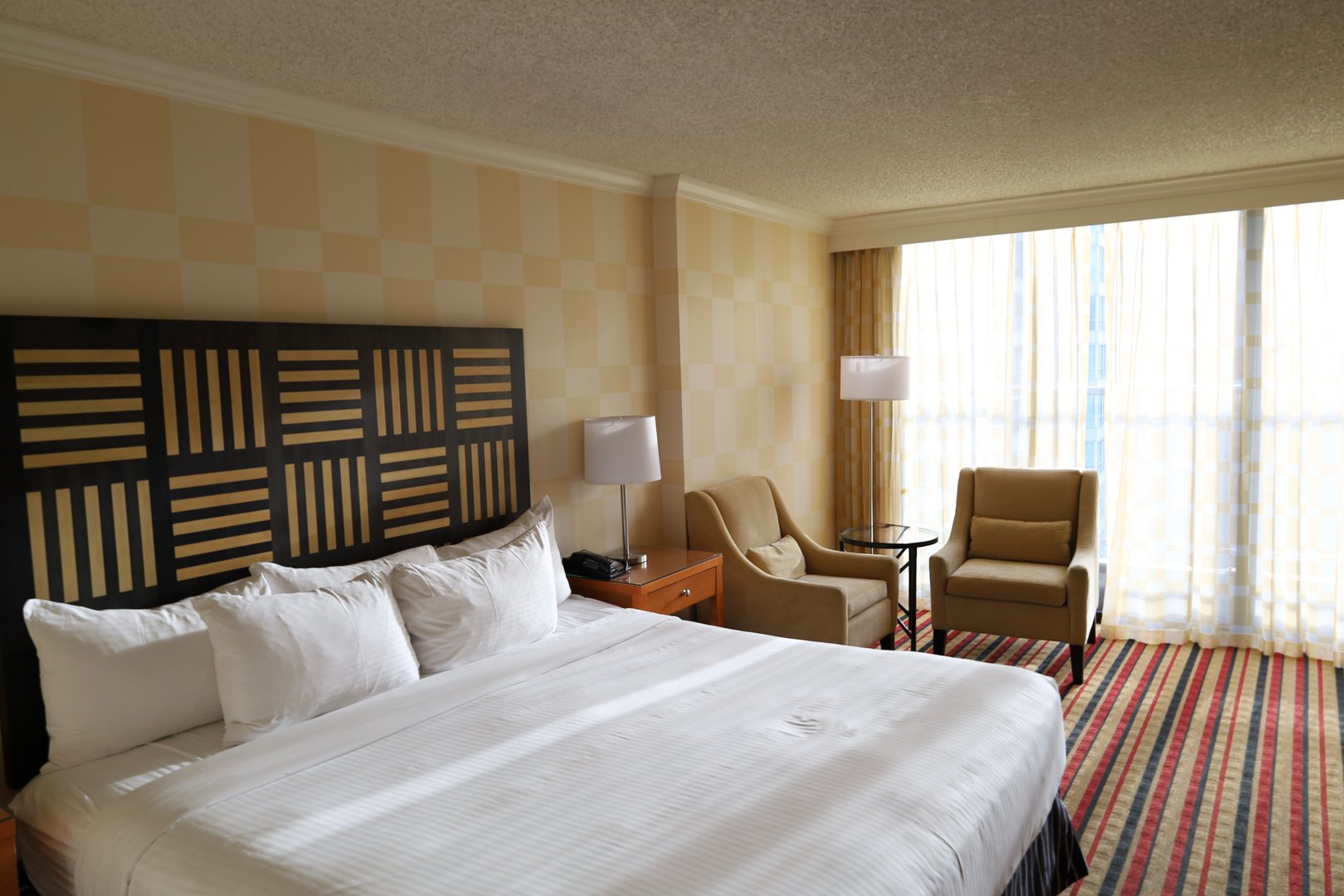 spacious room with a large bed, cozy armchairs, bedside lamps, and a panoramic window with sheer curtains, decorated in warm tones.