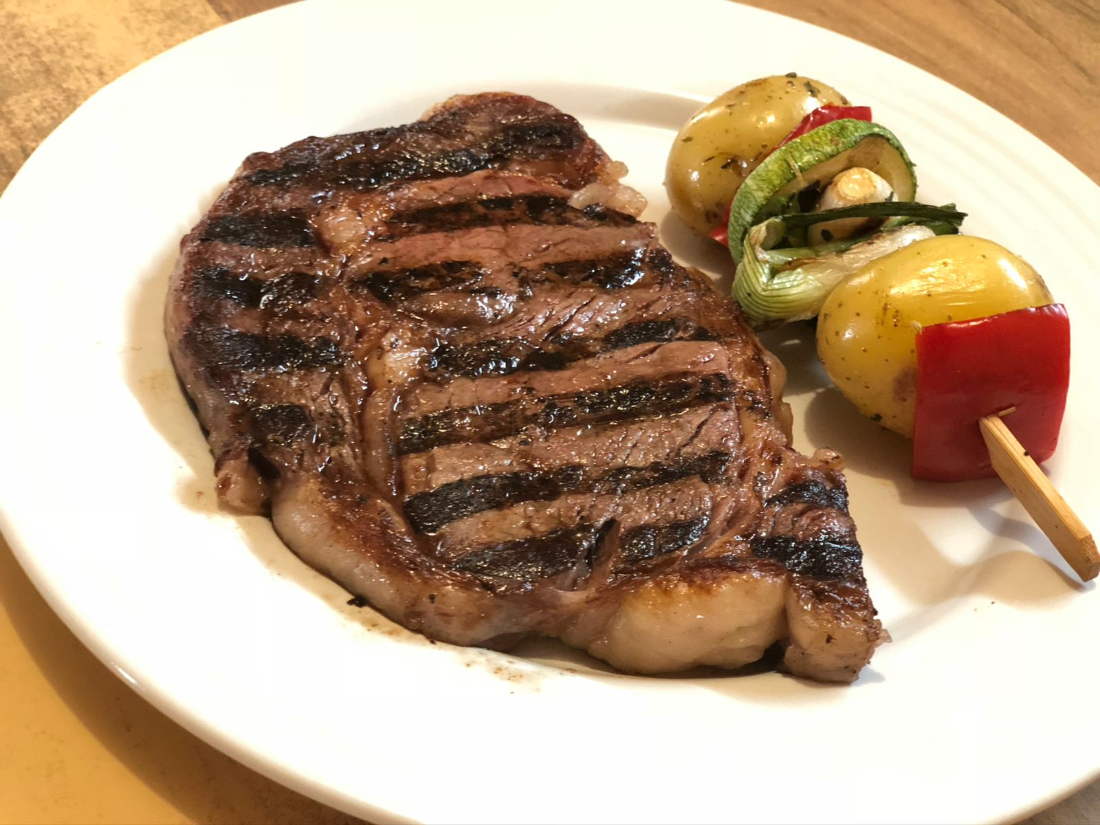 Grilled steak with a skewer of potatoes, peppers, and mushrooms on a white plate.