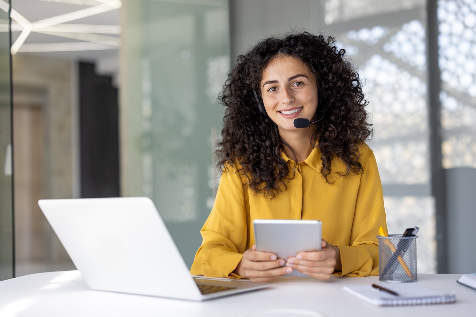 A woman with a headset smiles warmly while holding a tablet, ready to help customers with their inquiries