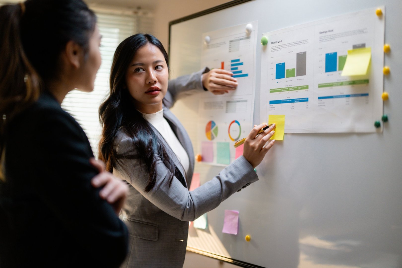 Asian businesswomen reviewing financial charts, collaborating on whiteboard, sharing strategic insights during workplace meeting