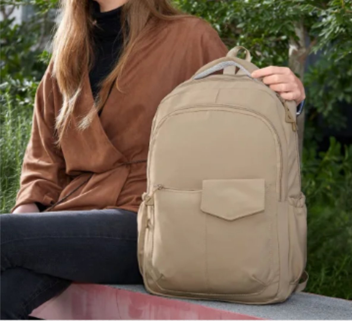 Person sitting on a bench holding a beige backpack, wearing a brown jacket and black pants, with greenery in the background.