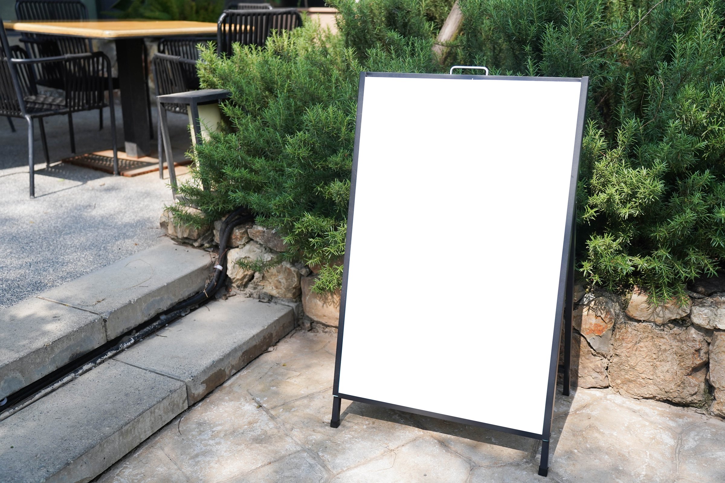 Blank white outdoor advertising stand sandwich board mock up template. Clear street signage board placed by an outdoor dinning area of a restaurant.
