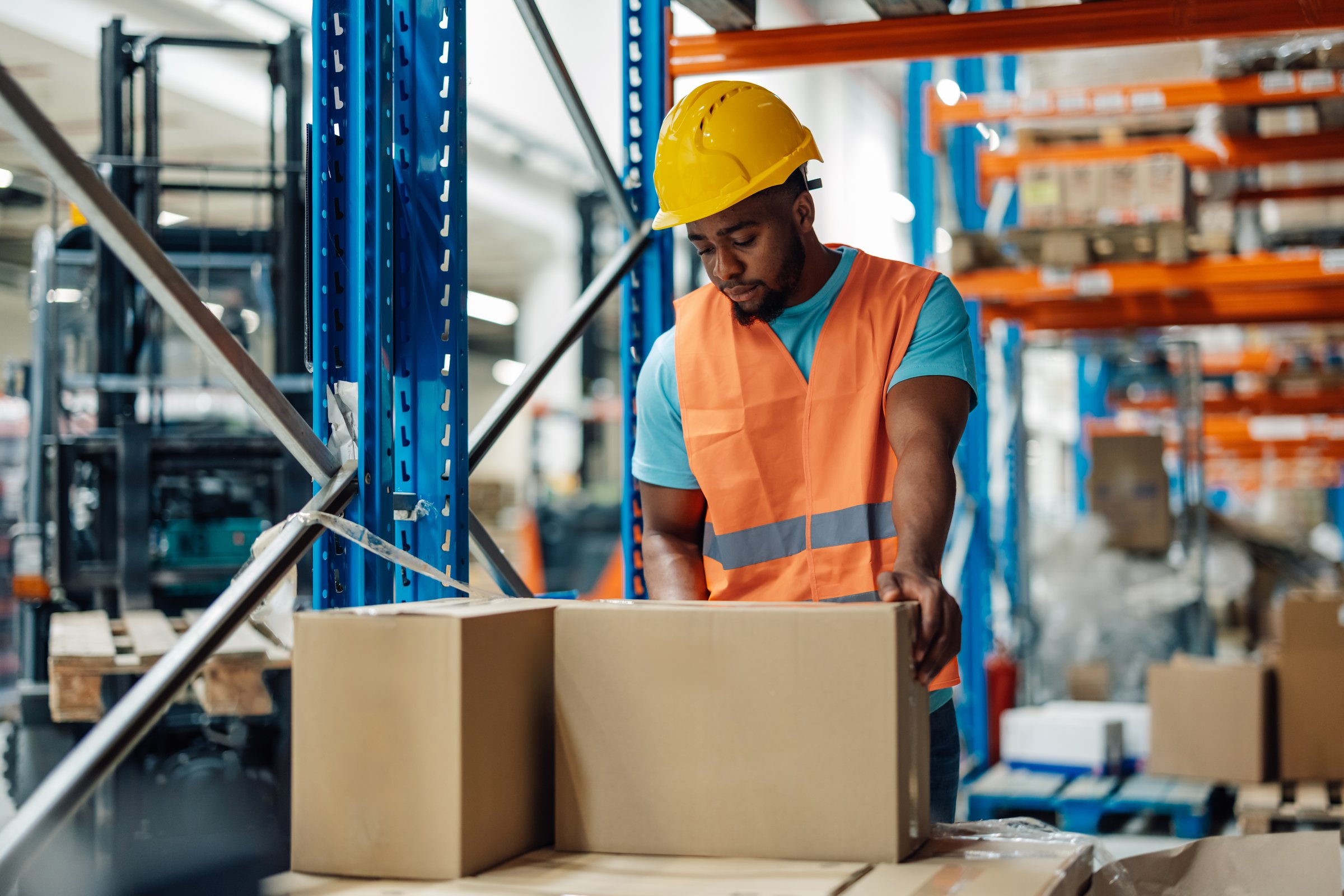African american warehouse worker is seen handling boxes in a large storage area, equipped with shelves and racks, the worker is dressed in a hard hat and safety vest, ensuring orderliness.