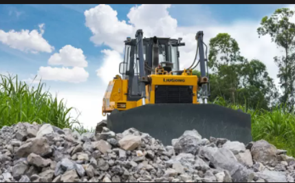 Yellow bulldozer pushing rocks in a grassy field with a clear blue sky and clouds in the background.