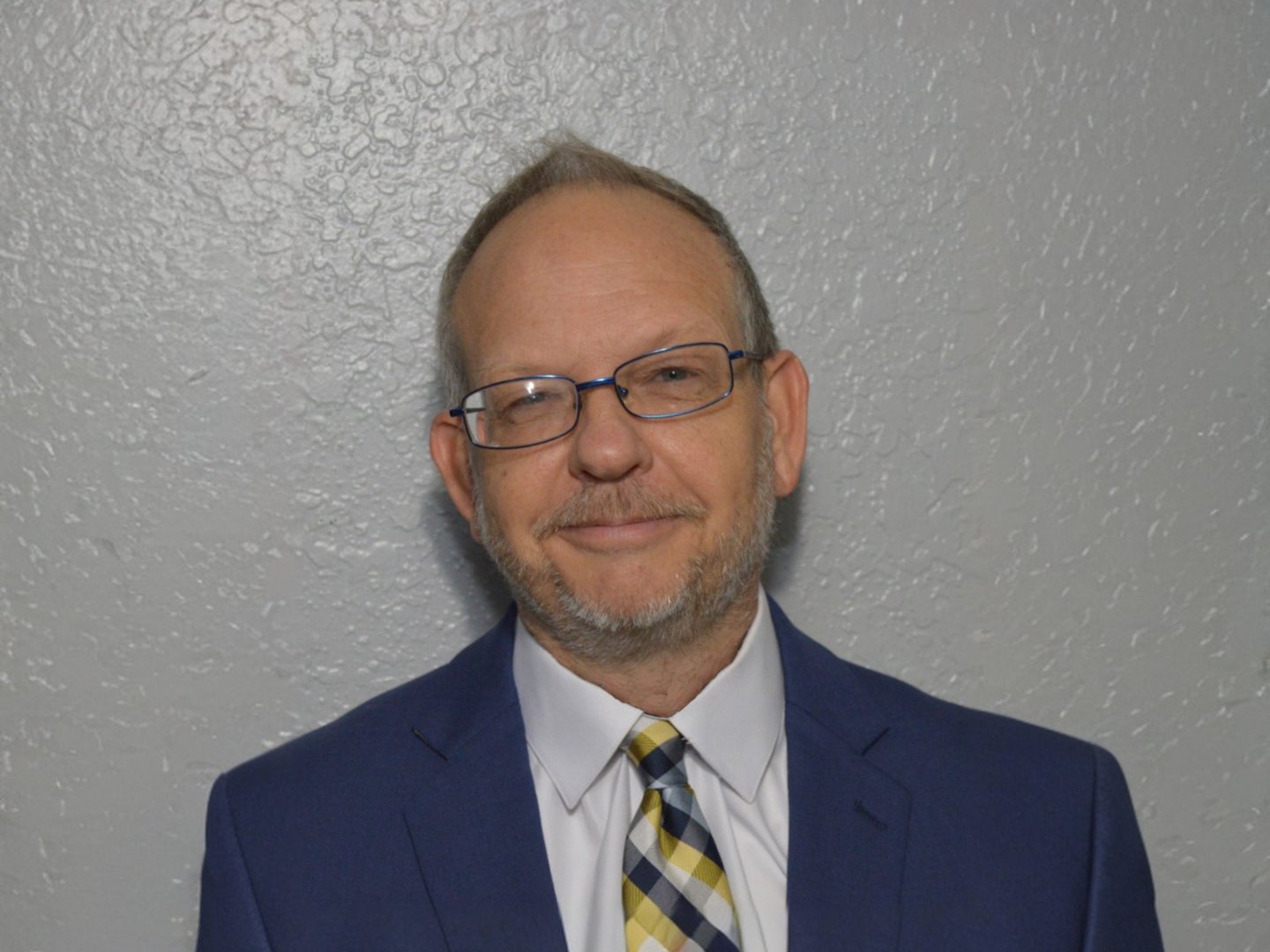 Man in glasses wearing a blue suit and patterned tie, standing against a textured gray wall, smiling slightly.
