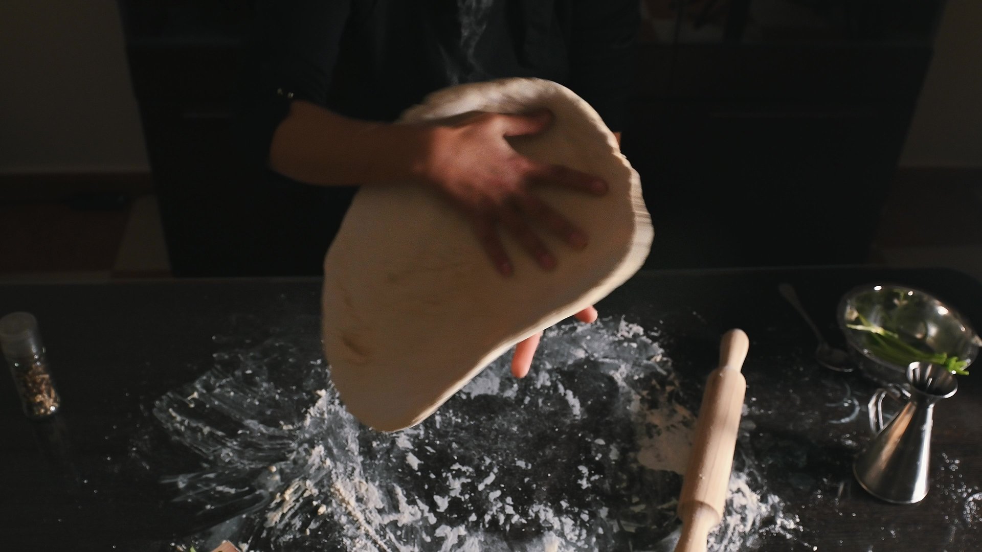 A scene capturing a person shaping pizza dough on a countertop sprinkled with flour, surrounded by various cooking tools and ingredients. Image highlights culinary preparation and creativity in the kitchen.