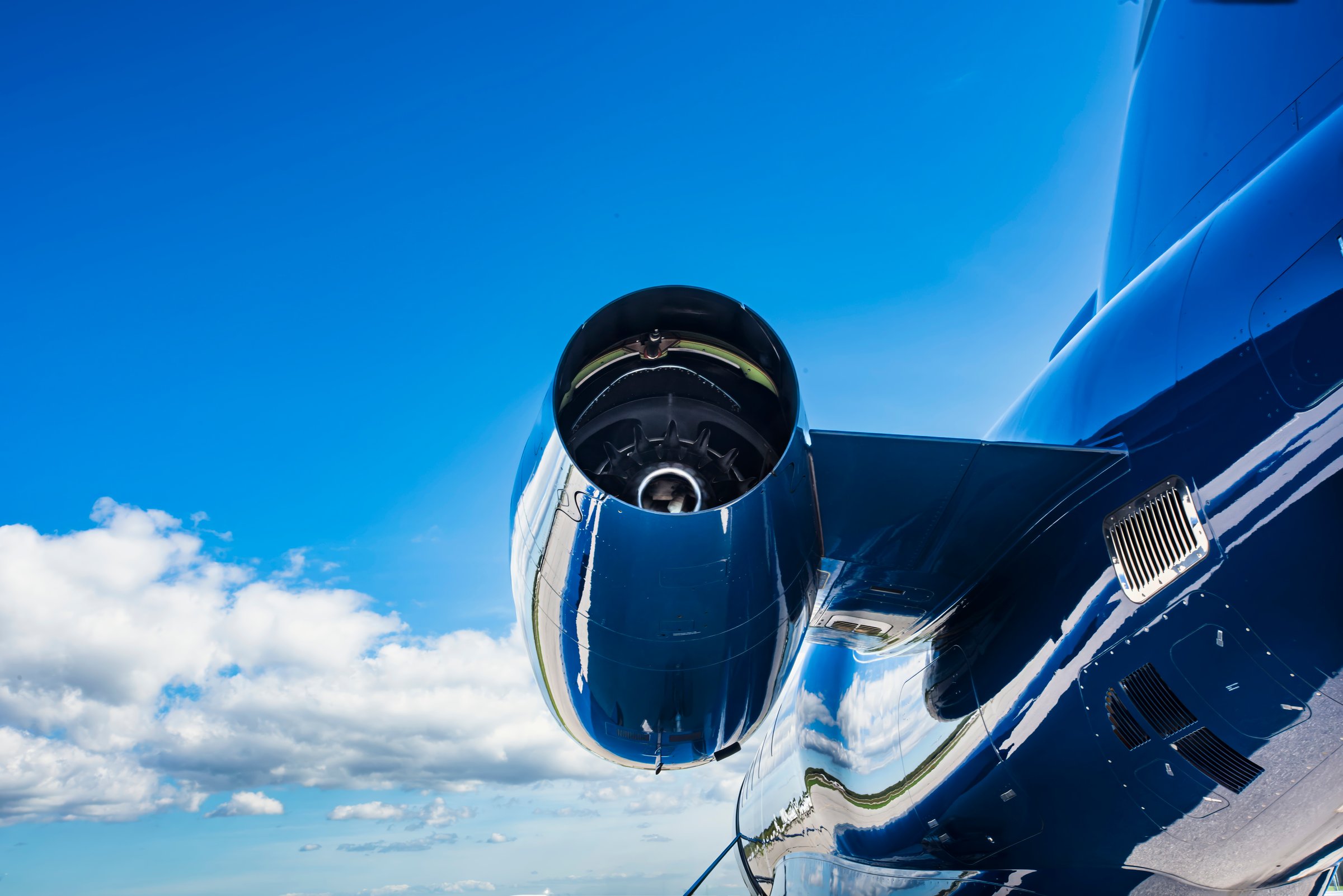 Close-up, low angle, rear view of a polished jet engine, fuselage, and part of the tail, with reflection detail and clouds beneath a vivid blue sky.