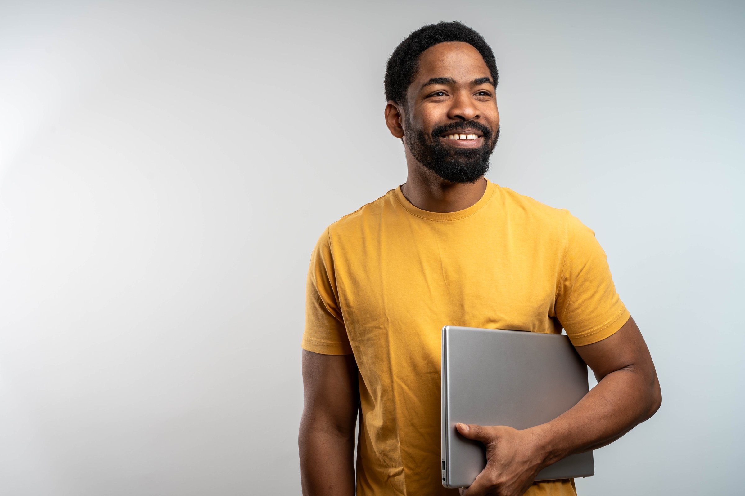 Smiling Young Handsome African Man Holding Closed Laptop on Bright Isolated Background in Yellow T-Shirt