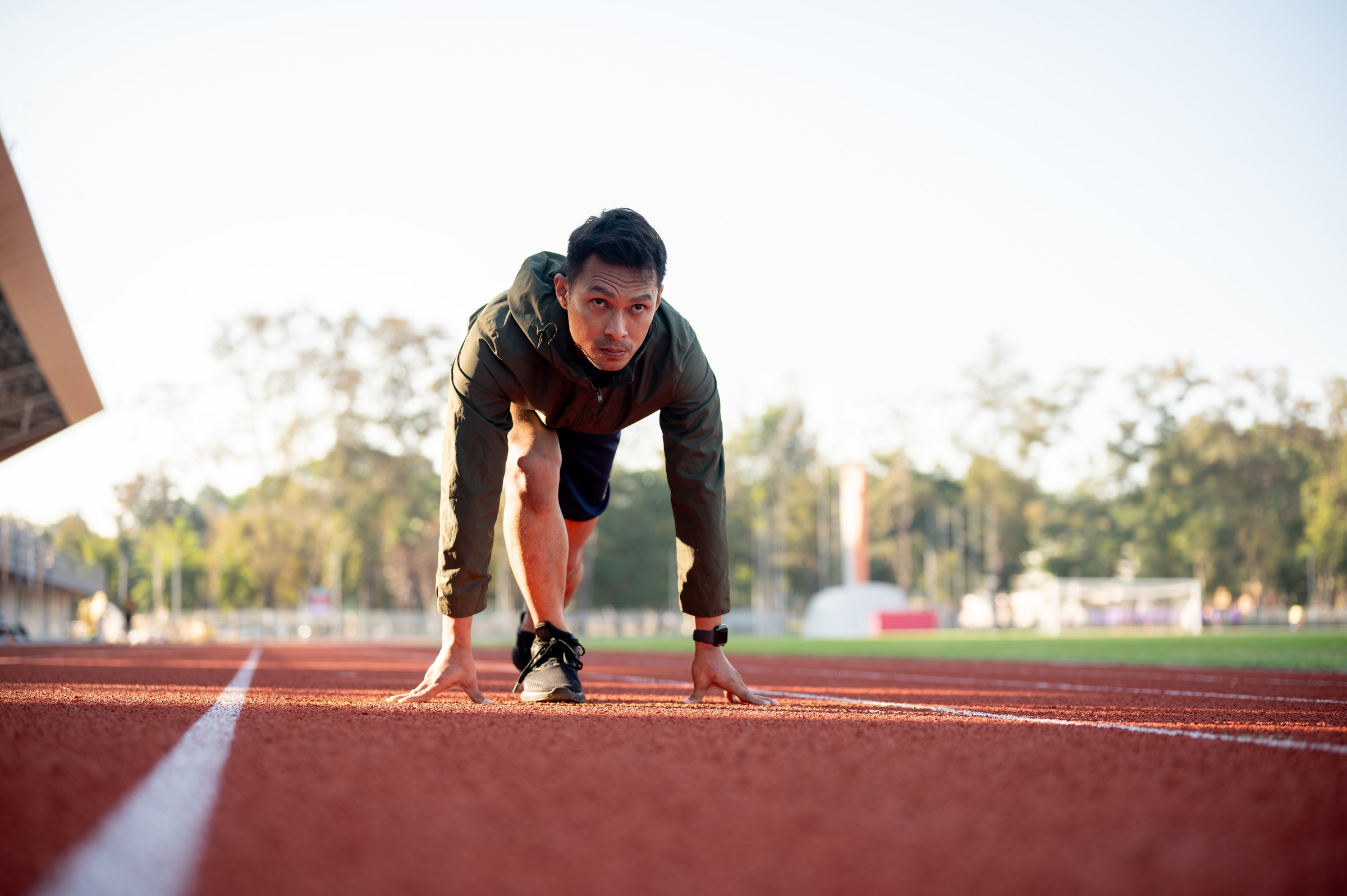 A strong Asian athletic male runner in sportswear trains at a stadium in the morning, preparing to run and getting ready at the starting line.