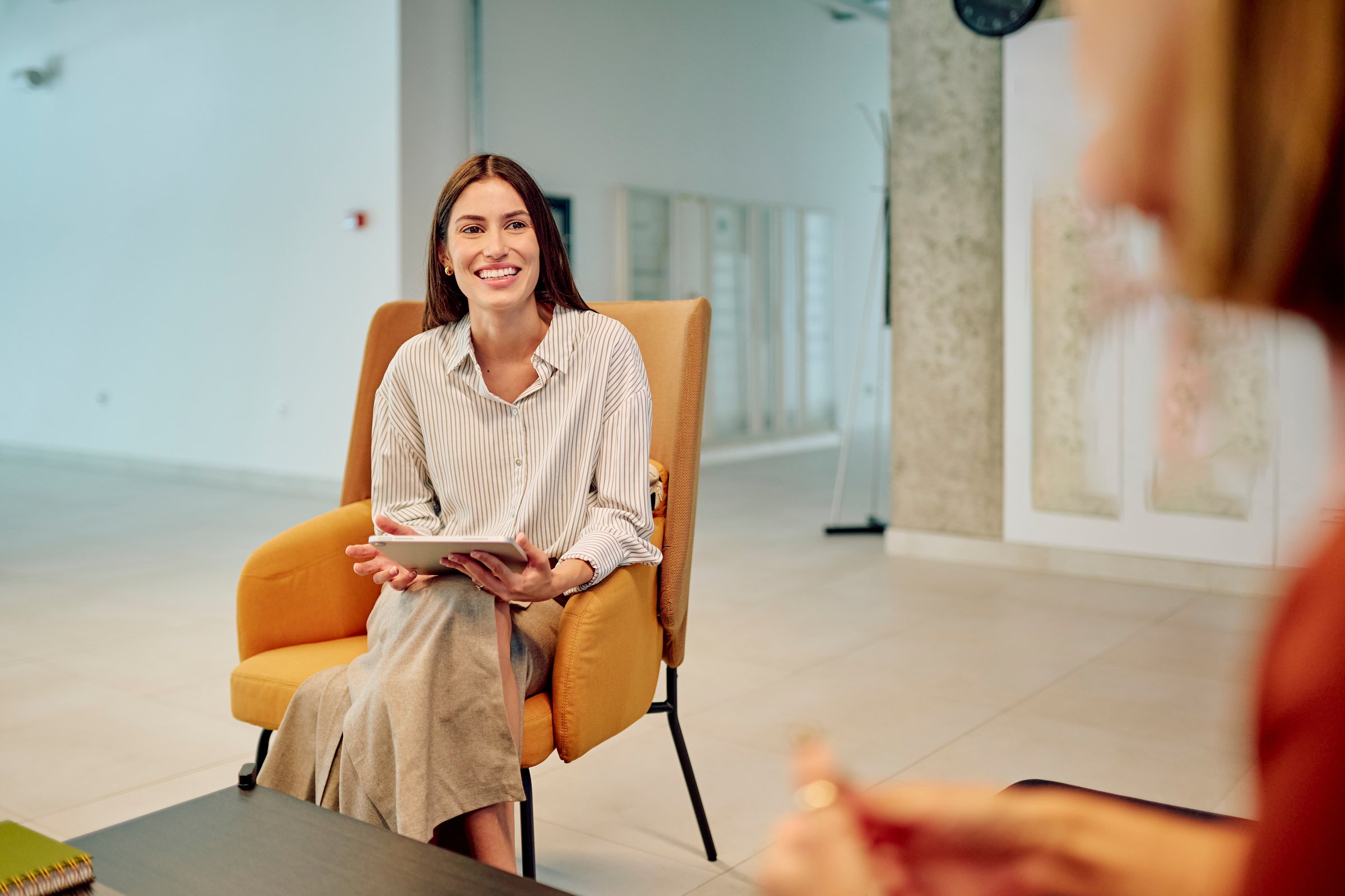 Businesswoman holding tablet and smiling while sitting on armchair and discussing with colleague in modern office