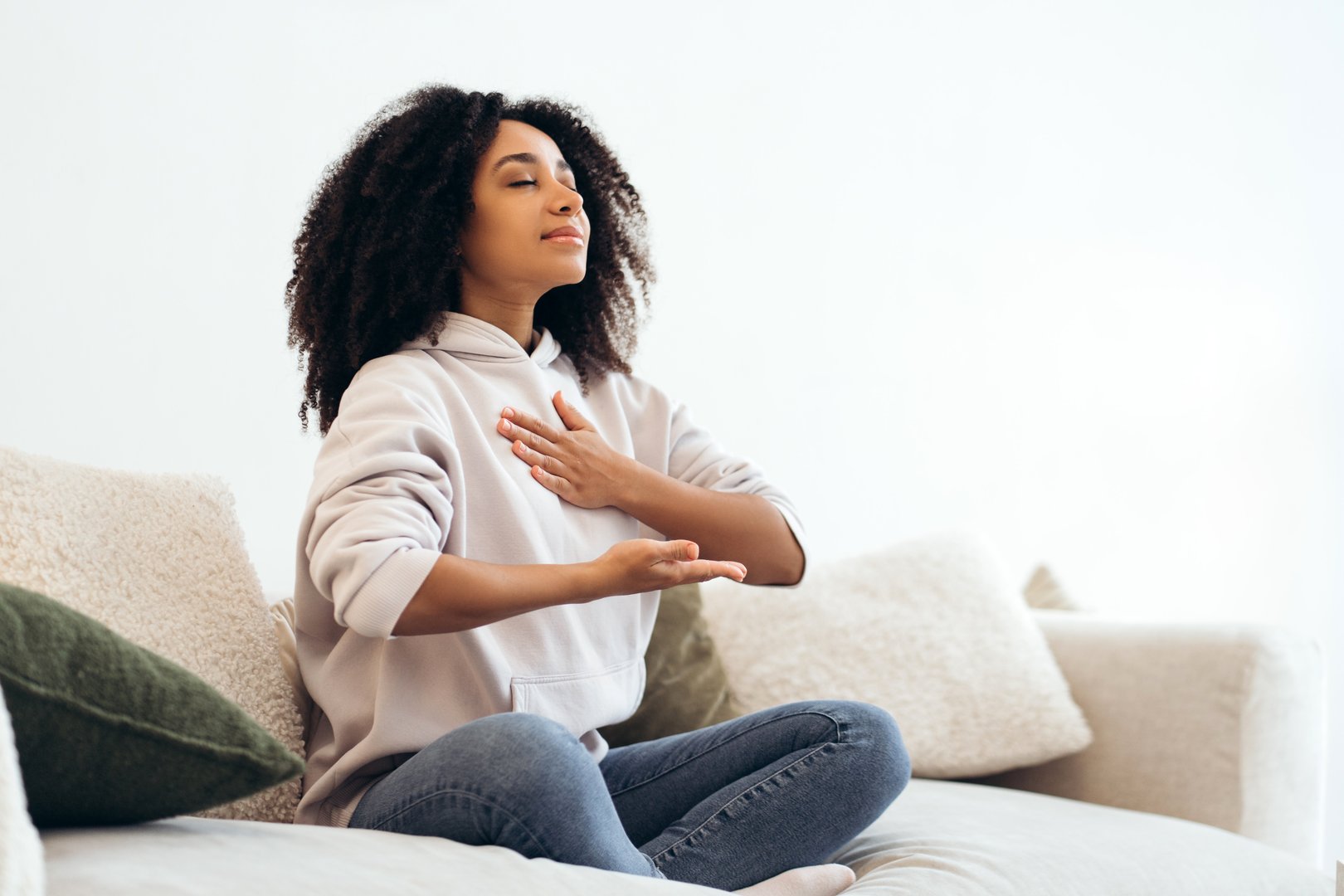 Serene young woman practicing breathing exercises while sitting in lotus position on comfortable sofa at home, enjoying meditation and relaxation