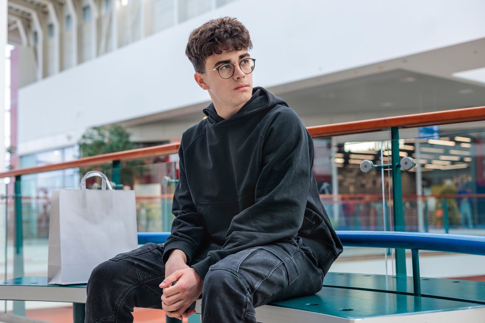 Teenage Boy in Stylish Black Hoodie Sitting in Shopping Mall with Smartphone and Shopping Bags – Modern Youth Lifestyle, Fashionable Streetwear, Digital Age, and Consumer Culture Concept. Copy space
