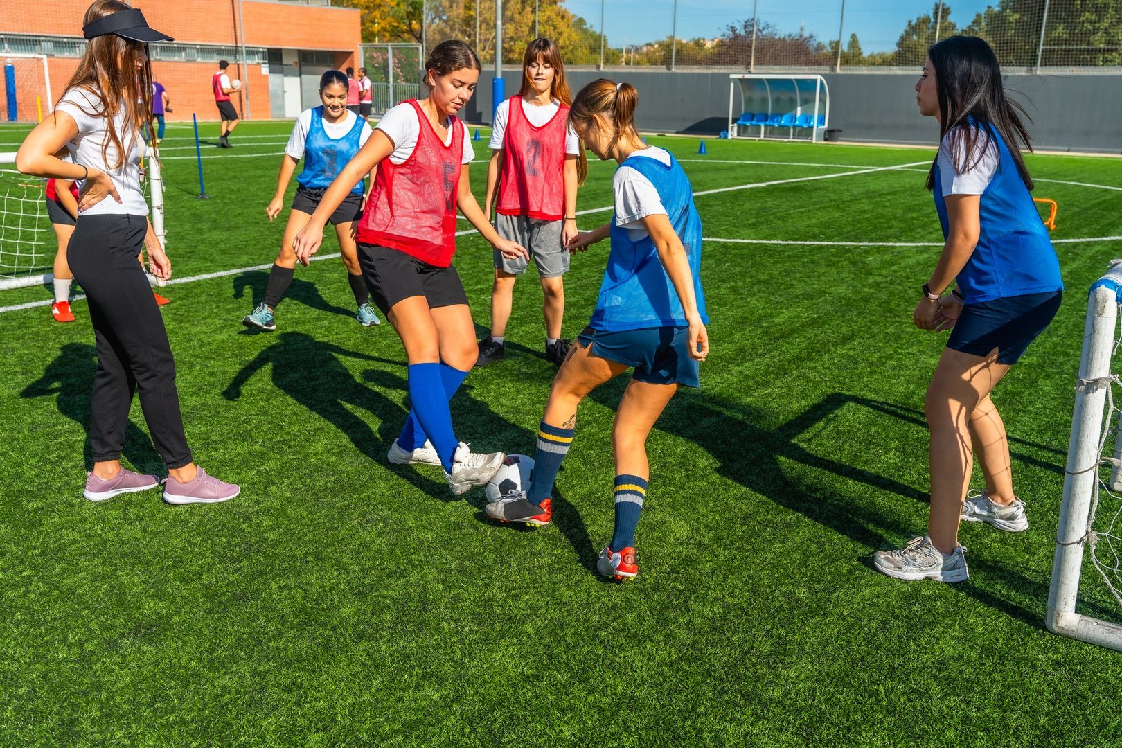 Teenagers participating in a girls' soccer training session, actively playing on a vibrant green artificial turf field with a coach overseeing the practice