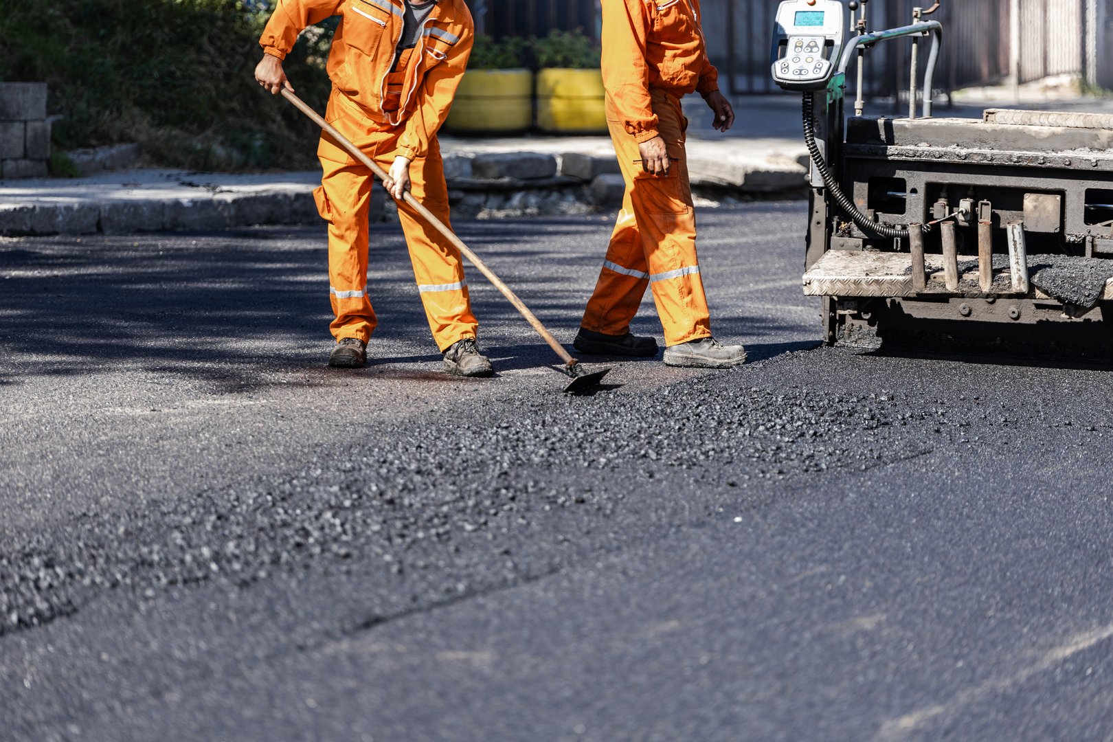 Two workers in bright orange uniforms are evenly spreading asphalt on a freshly paved road under clear skies during the daytime.