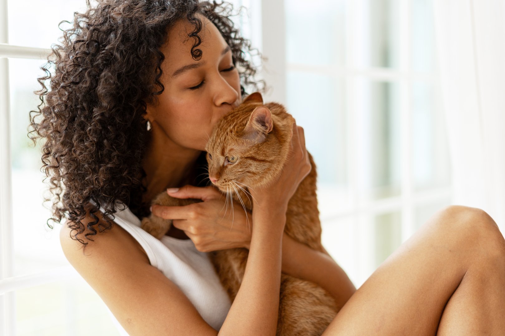 Young adult woman lovingly holding and kissing her ginger cat by a window. Captures the bond and affection between humans and their pets in a serene setting