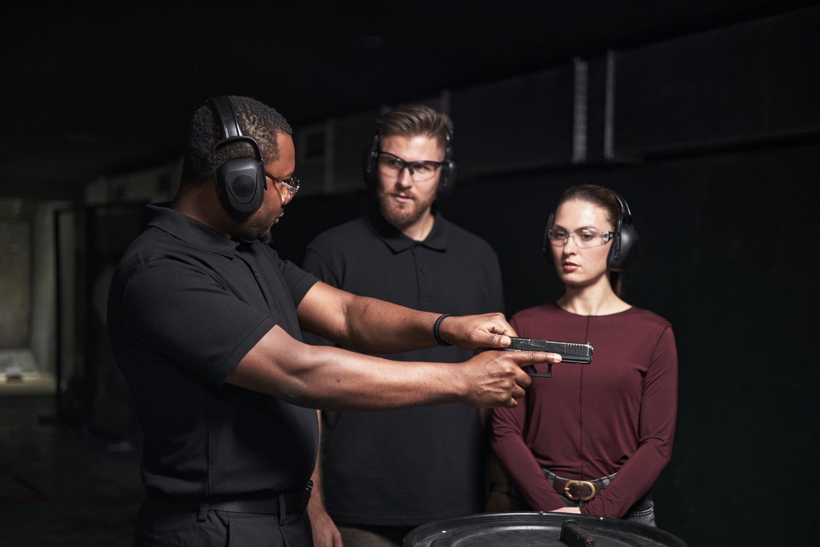 African American man instructing Caucasian young adult man and woman at indoor shooting club, all wearing protective earmuffs and safety glasses, focusing on handgun handling