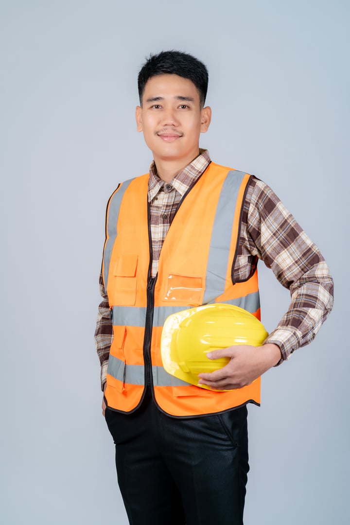 Young Asian male engineer or worker wearing yellow hard hat and reflective vest standing on grey background.
