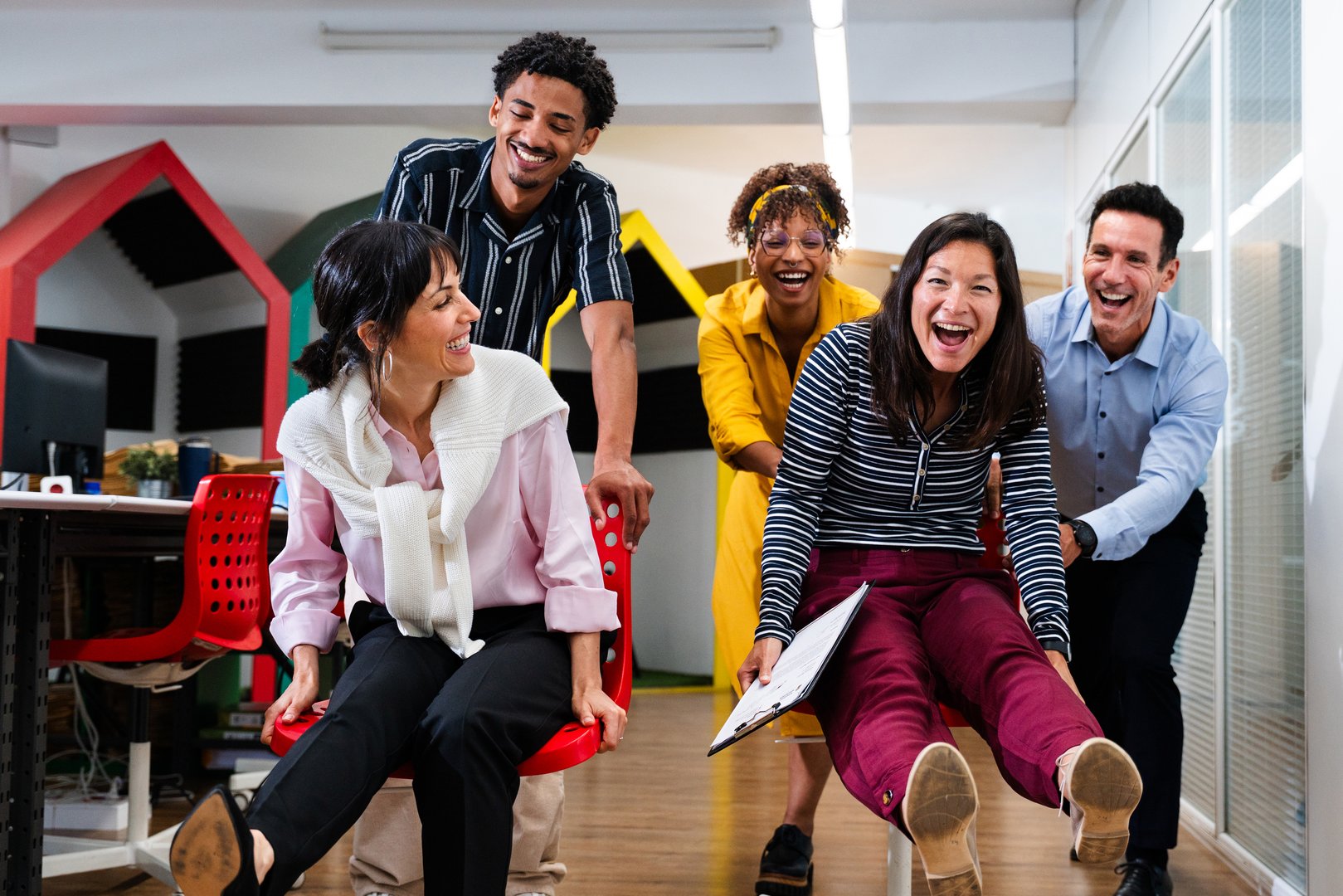 Multi-ethnic businesspeople are enjoying a playful moment, racing on office chairs through a vibrant and modern startup office space, showcasing teamwork and positive energy